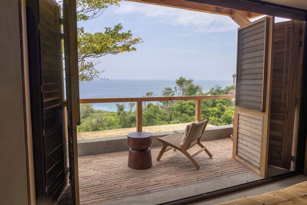 An open balcony with a wooden floor, a round table, and a chair, overlooking a scenic view of the sea and greenery.