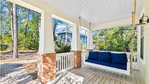 A covered porch with a swing seat, brick columns, and a view of surrounding trees and a house.