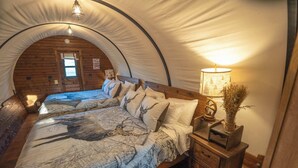 A cozy tented room with a bed featuring a deer print bedspread, a bedside lamp, and a small wooden nightstand. - Modern_ Unique_Old West Wagon_Spacious_Views (Chilton)