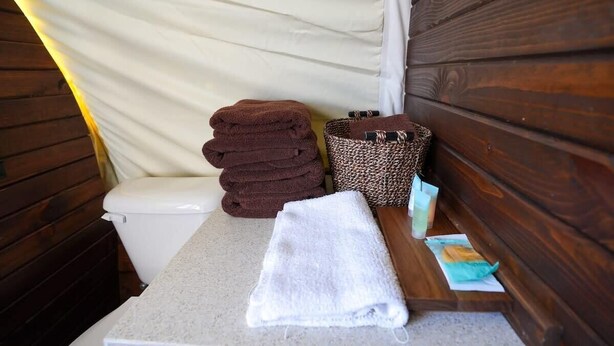 A small bathroom with a wooden bench, a white toilet, and a stack of brown towels. - Modern_ Unique_Old West Wagon_Spacious_Views (Chilton)