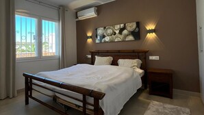 Une chambre avec un lit en bois, un luminaire au plafond, un climatiseur fixé au mur et une fenêtre avec des rideaux.