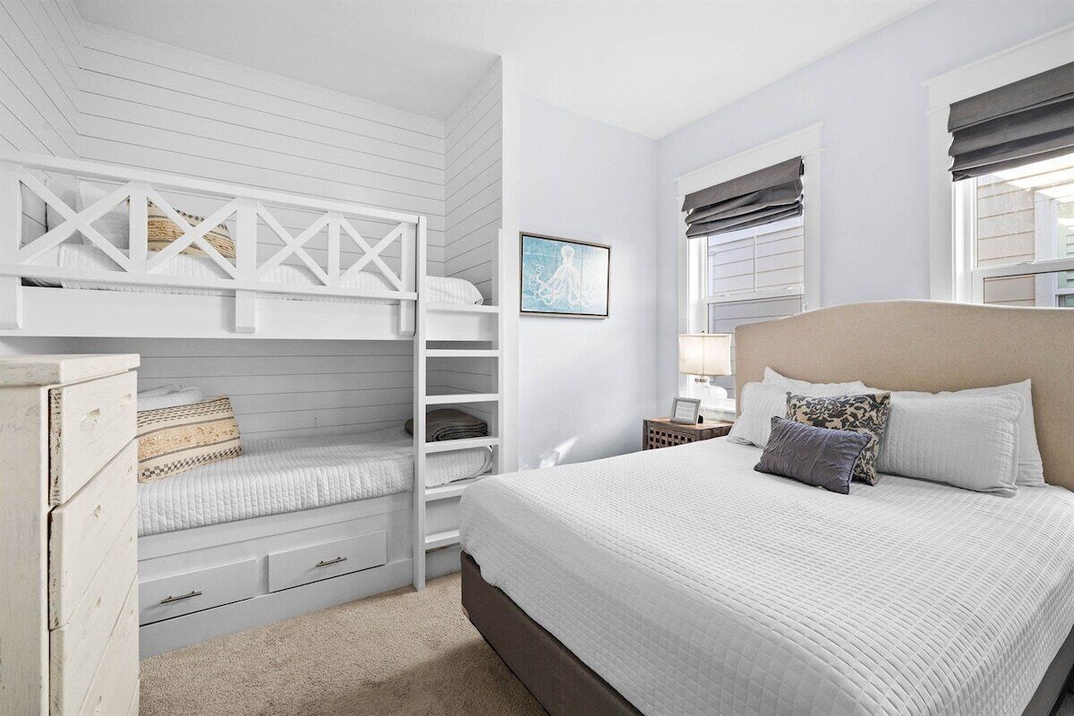 A bedroom with a bunk bed, a large bed with pillows, a nightstand, a lamp, and a framed picture on the wall.