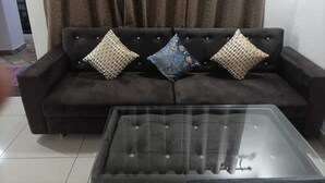 A dark-colored sofa with three patterned cushions, a glass-top coffee table, and a curtain in the background. - Maa Bhawani Homestay (Reasi)