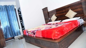 A modern bedroom with a wooden headboard, a large bed with a red and floral bedspread, and a window with blue patterned curtains. - Maa Bhawani Homestay (Reasi)