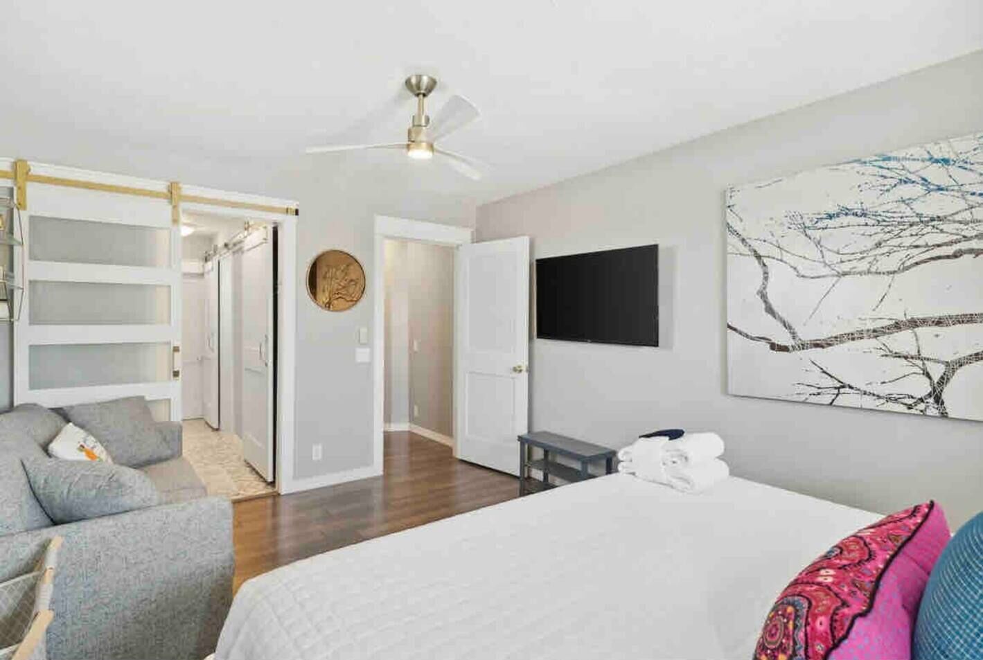 Ein modernes Schlafzimmer mit einem Bett, einem grauen Sofa, einem Deckenventilator, einem Fernseher an der Wand und einem großen abstrakten Gemälde.