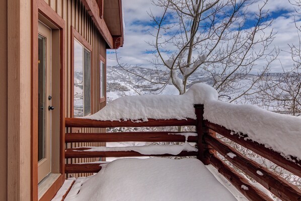 A wooden balcony with snow accumulation, a wooden door, and a view of a snowy landscape with trees and mountains.