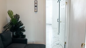 Een woonkamer met een donkergrijze modulaire bank, een blauwgroen poef en een grote groene plant. Een ingelijste foto hangt aan de muur. De kamer heeft houten vloeren en een witte deur die leidt naar een andere kamer.
