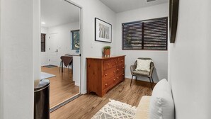 Une pièce avec un plancher en bois, un canapé blanc, une commode en bois, un tableau encadré au mur et une fenêtre avec des stores.