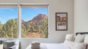 A hotel room with a bed, a chair, a window with a mountain view, and a framed picture on the wall.