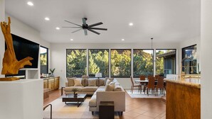 Ein modernes Wohnzimmer mit einer Couch, einem Couchtisch und einem Fernseher. Große Fenster, ein Deckenventilator und eine Küchenzeile mit Esstisch und Stühlen sind ebenfalls vorhanden.