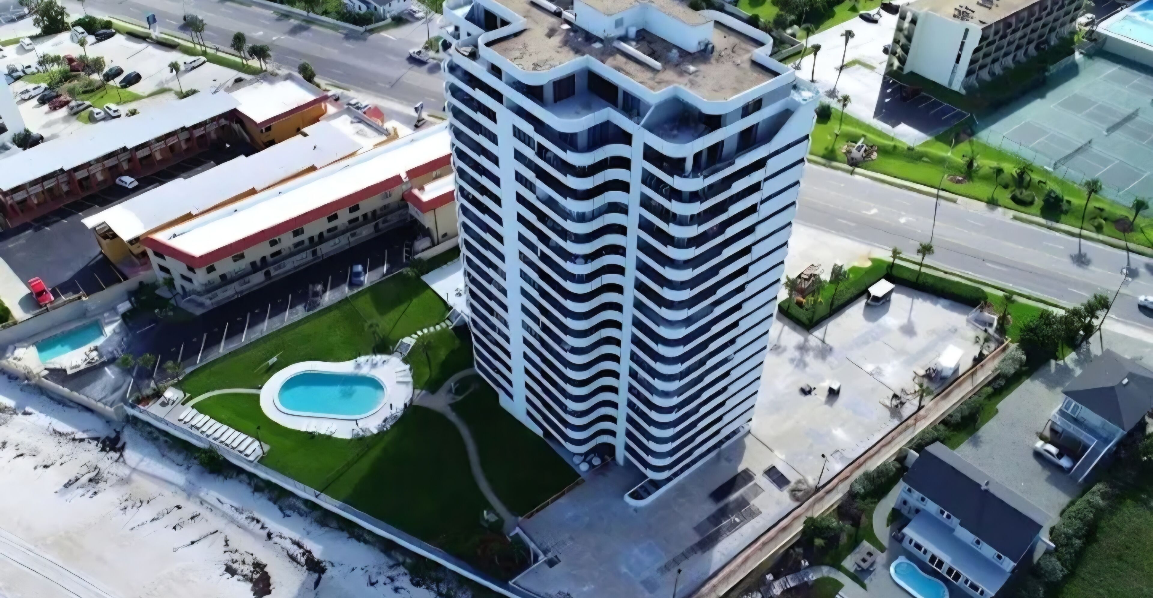 A high-rise building with a rooftop pool, surrounded by green lawns and a parking area.