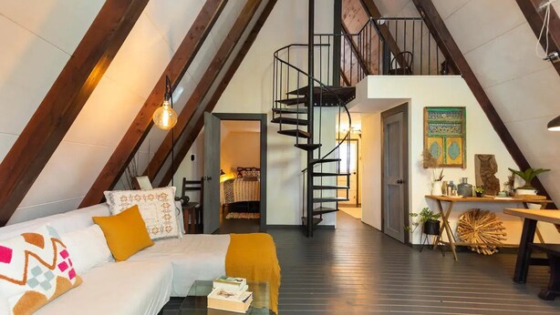 A loft-style room with a high ceiling, exposed wooden beams, a spiral staircase, a white sofa with colorful cushions, a glass coffee table, and a small dining area with a wooden table and chairs.