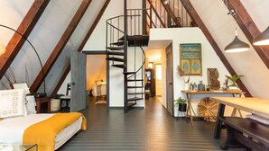 A loft-style bedroom with a spiral staircase, a bed with a yellow throw, and a dining area with a wooden table and chairs.