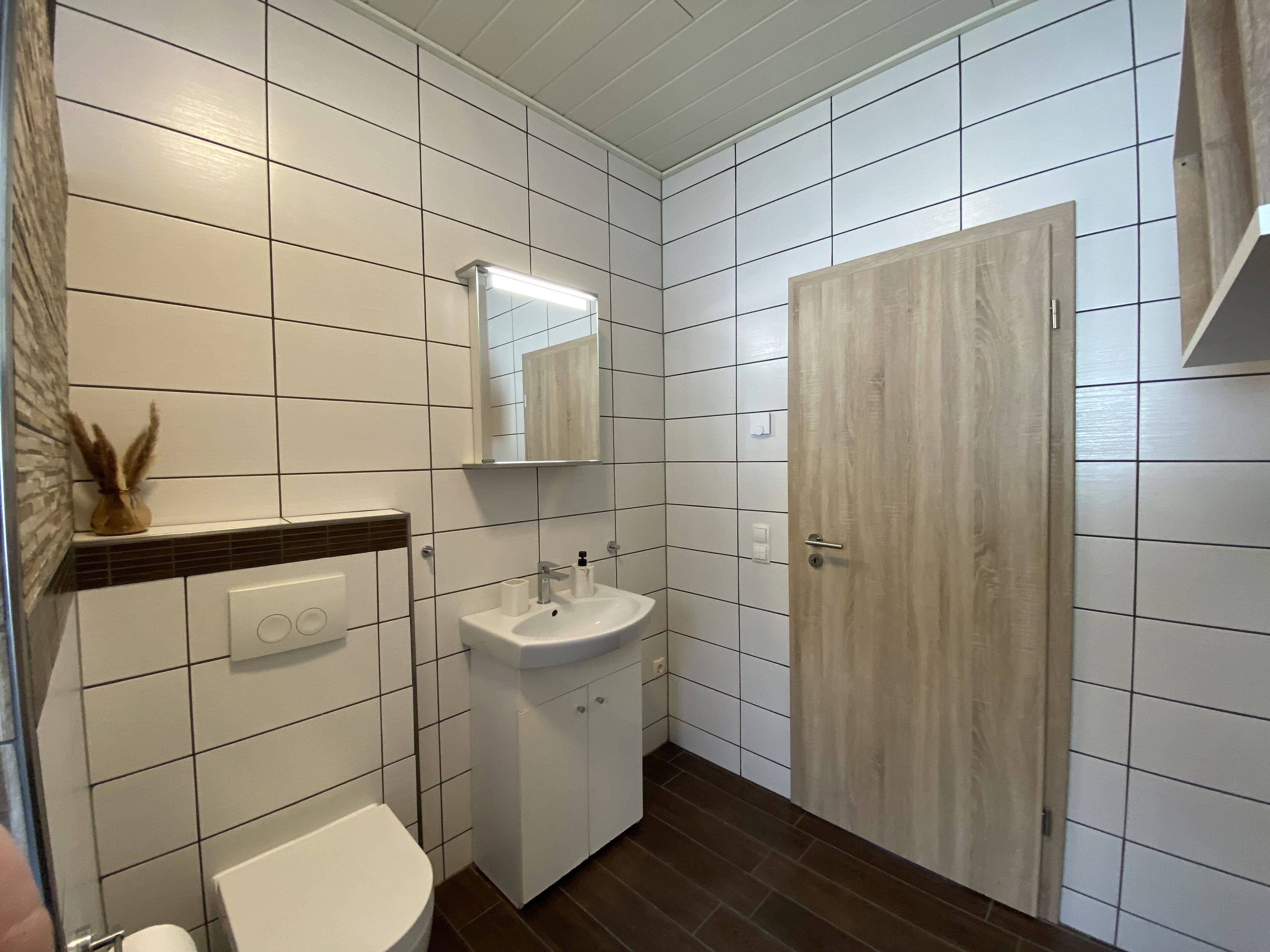 Un baño con paredes de azulejos blancos, una puerta de madera, un lavabo blanco y un inodoro.