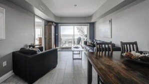 Una sala moderna con un sofá oscuro, una mesa de comedor de madera y vistas a un balcón exterior a través de un amplio ventanal corredero.