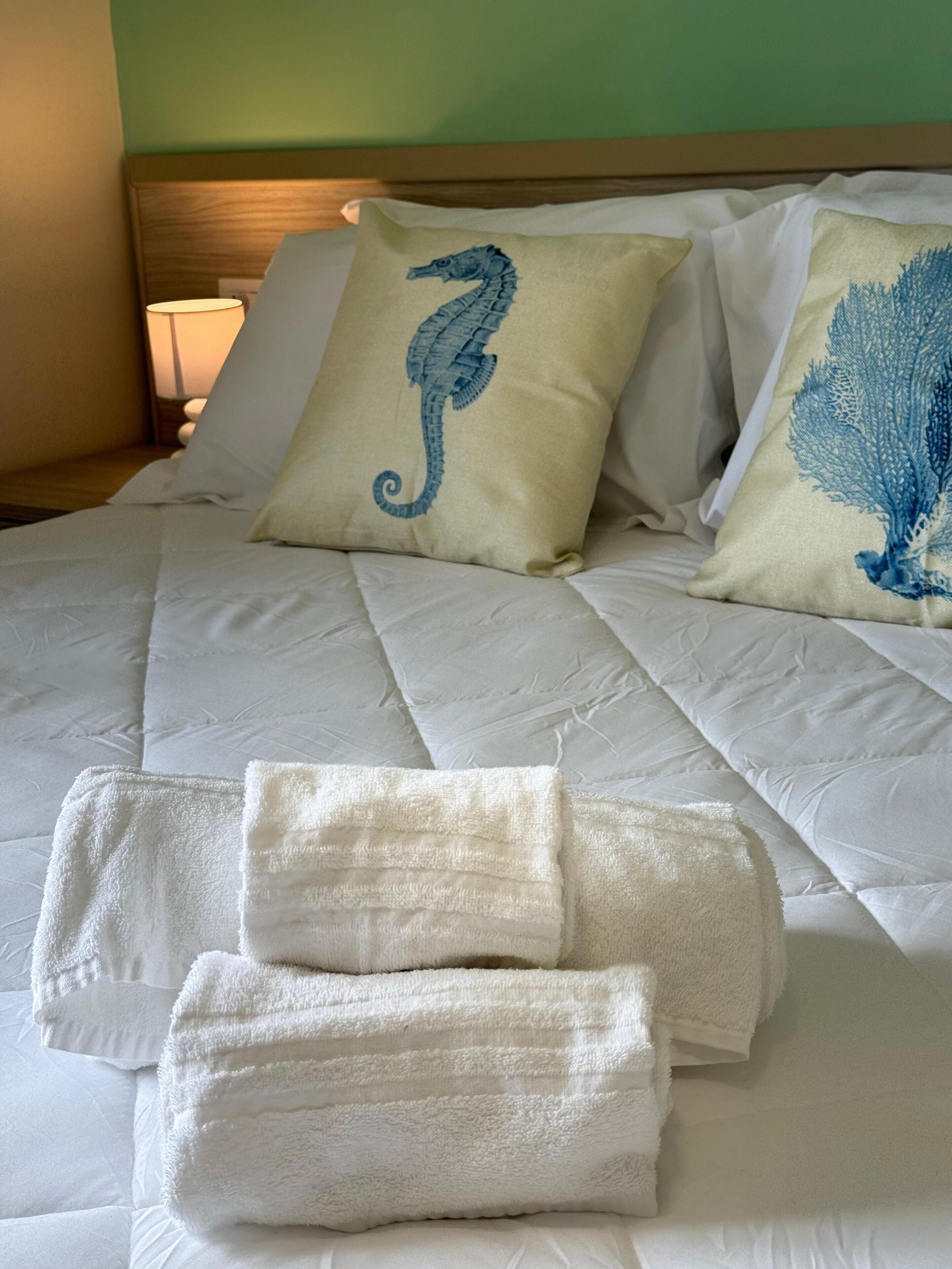Una cama bien hecha con ropa de cama blanca y dos almohadones decorativos, uno con diseño de caballito de mar y otro con motivos de coral azul.