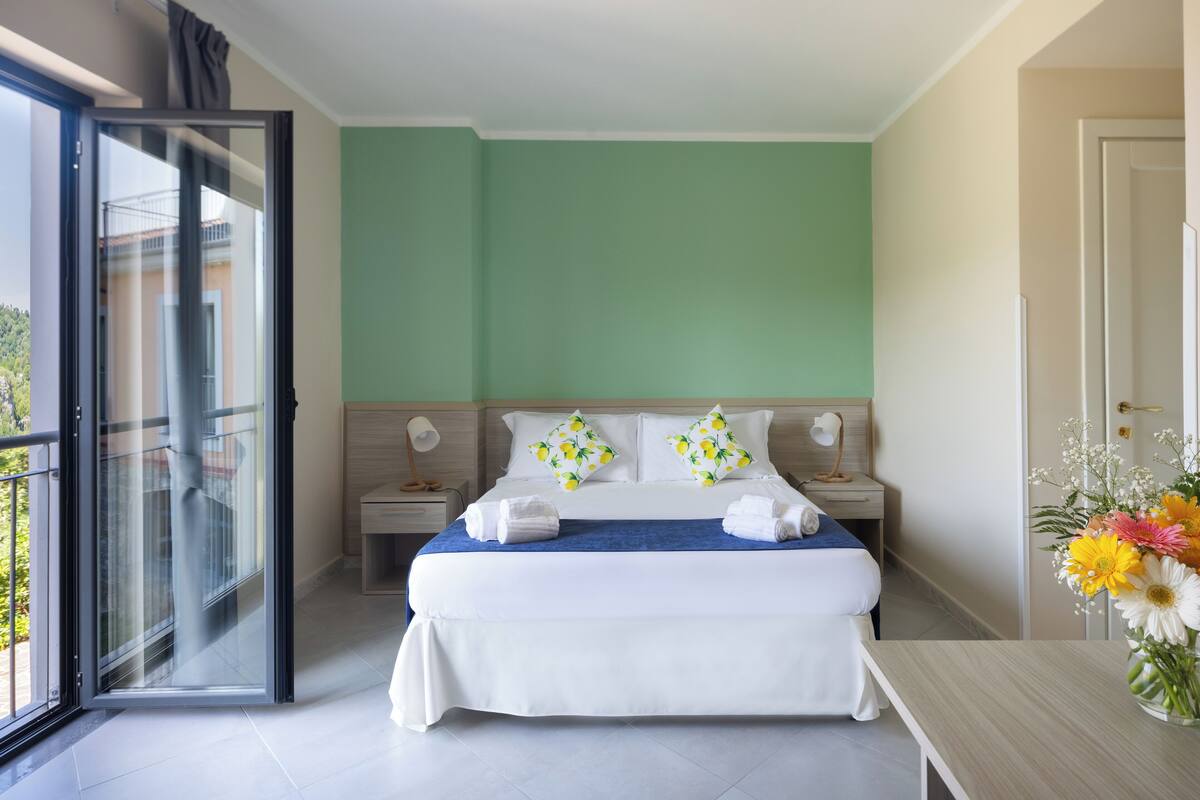 Une chambre d’hôtel avec un grand lit, une table de chevet, un bureau et une porte-fenêtre coulissante donnant sur un balcon.