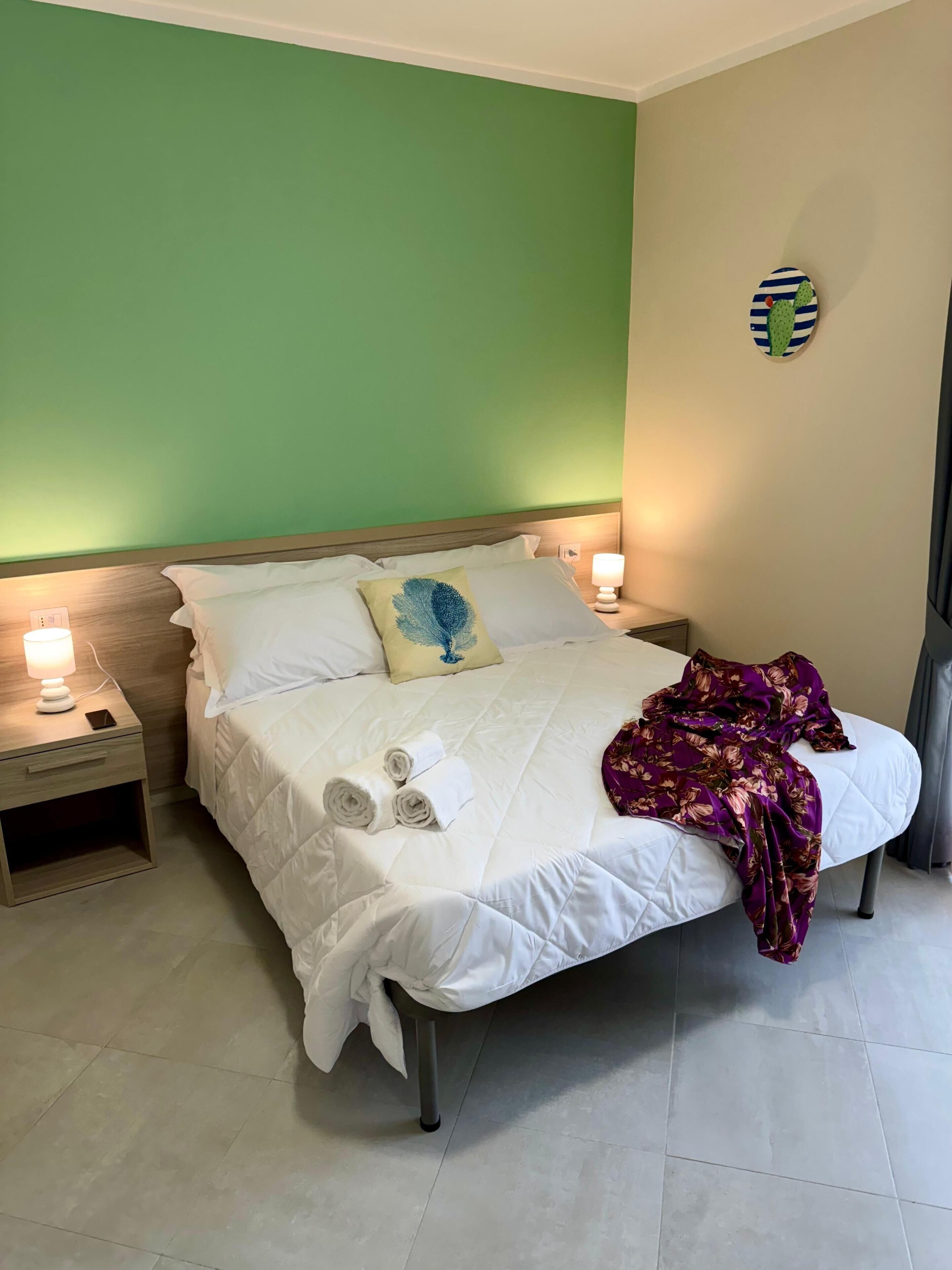Un lit bien fait, avec des draps blancs et une couverture à motifs floraux violets, deux serviettes enroulées et un coussin décoratif orné d’un motif de plumes bleues.