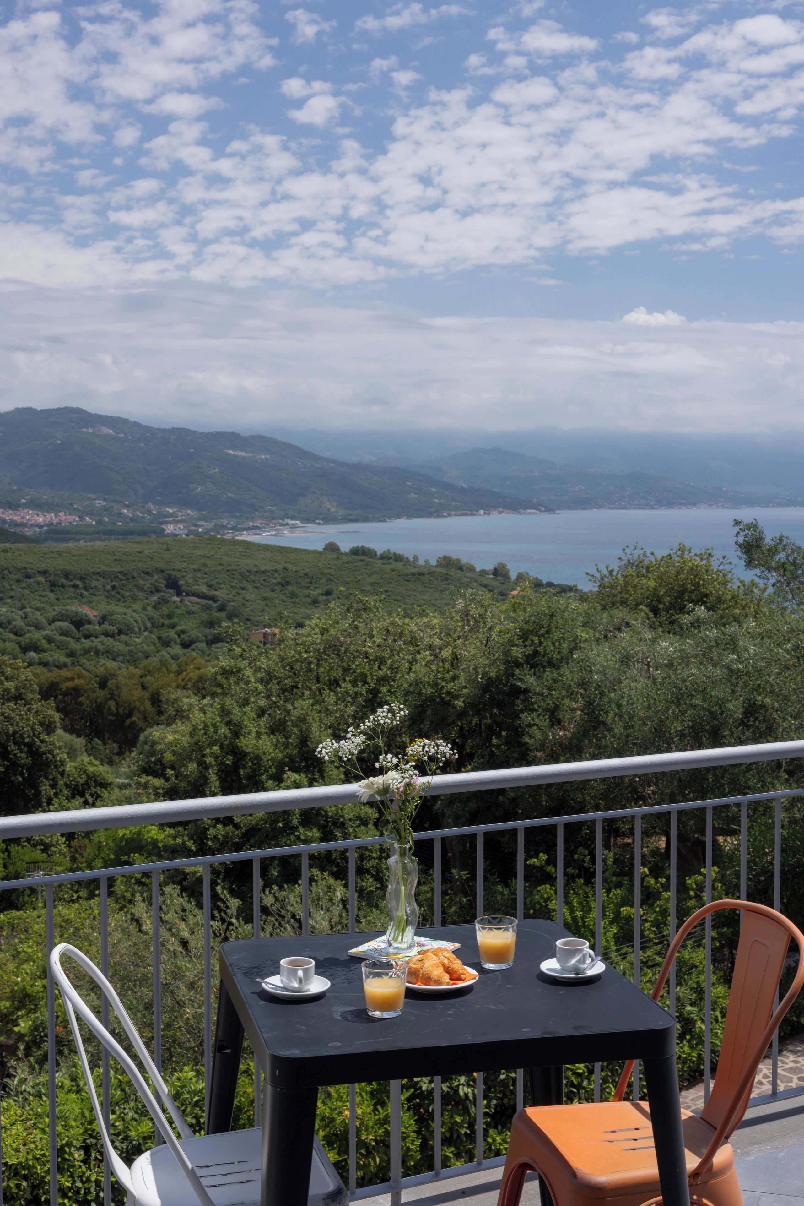 Una mesa con una bebida, un plato de comida y un jarrón de flores en un balcón con vistas panorámicas a la costa y colinas.