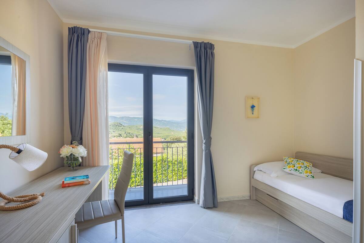 Une chambre à coucher comprenant un lit, un bureau, une chaise et donnant sur un paysage verdoyant grâce à une grande fenêtre.