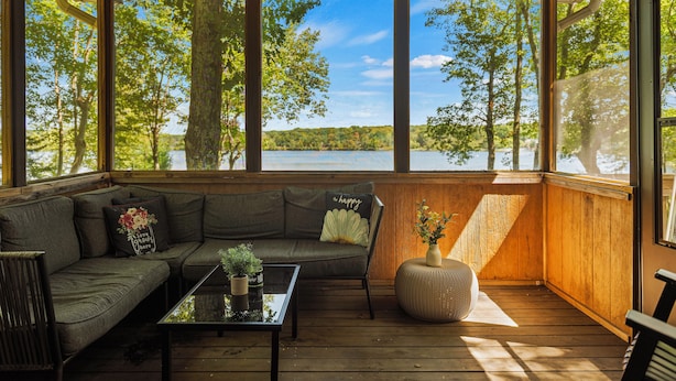 A cozy cabin with a wooden deck, a sofa, a coffee table, and a view of a lake surrounded by trees.