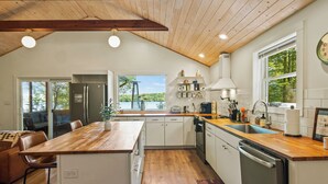 A modern kitchen with wooden countertops, white cabinets, and a large island. There is a view of a lake through the window, and the kitchen is equipped with a dishwasher and a sink.