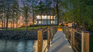 Un pont en bois menant à une maison dotée de nombreuses fenêtres, entourée d’arbres et bordée par une rivière.