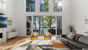Un salon spacieux avec un grand canapé, deux fauteuils et une table basse. Il y a un téléviseur sur le côté gauche et une grande fenêtre donnant sur des arbres et un plan d’eau.