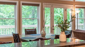 A dining area with a table, chairs, a vase with branches, and large windows with a view of greenery.