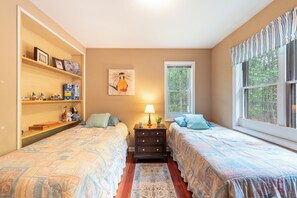 A bedroom with two beds, a nightstand with a lamp, a shelf with various items, and a window with a view of trees.