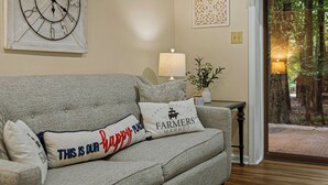 A living room with a sofa, decorative pillows, a wall clock, a table lamp, and a view through a glass door. - Frolic on Fern by AvantStay | Enclosed Deck w/ Forest Views, Bar (Blakeslee)
