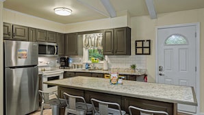 A modern kitchen with stainless steel appliances, a large island with bar stools, and a decorative window above the sink. - Frolic on Fern by AvantStay | Enclosed Deck w/ Forest Views, Bar (Blakeslee)