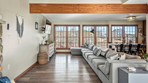 A spacious living room with a large sectional sofa, wooden beams on the ceiling, and large windows offering a view of the outdoors. - Laker's Pointe by AvantStay | Lakefront Private Peninsula | Hot Tub, Game Room (Long Pond)
