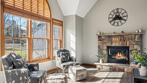 A living room with a fireplace, two armchairs, and a large window with blinds. - Laker's Pointe by AvantStay | Lakefront Private Peninsula | Hot Tub, Game Room (Long Pond)