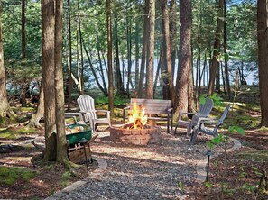 A forest campsite with a fire pit, chairs, and a bench.