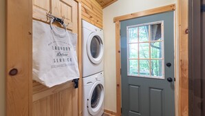 A laundry room with two stacked washing machines, a wooden door, and a window with a view of trees. - Arrowhead Lake Retreat by AvantStay | Shared Pool, Game Room, Firepit (Pocono Lake)