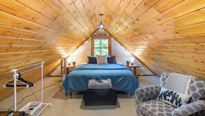 A cozy attic bedroom with a sloped ceiling, a bed with blue bedding, a chair with patterned cushions, and a small table with a lamp. - Arrowhead Lake Retreat by AvantStay | Shared Pool, Game Room, Firepit (Pocono Lake)