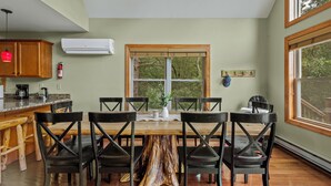 A dining area with a wooden table and chairs, a kitchen with a coffee maker, and a large window with a view of trees.
