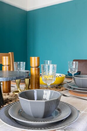 A dining table set with grey plates, bowls, and glasses, surrounded by wooden chairs and a blue wall.