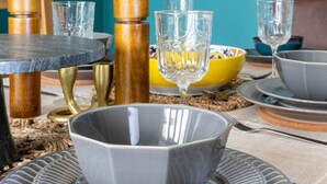Una mesa de comedor dispuesta con platos, tazones y vasos grises, rodeada de sillas de madera y una pared azul.