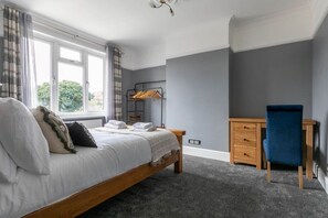 A bedroom with a bed, a desk, and a chair. There is a window with curtains, a radiator, and a wall-mounted coat rack.