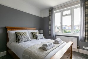 Una cama bien tendida con sábanas blancas y una manta gris, un cabecero de madera y una ventana con cortinas.