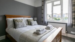 Una cama bien tendida con sábanas blancas y una manta gris, un cabecero de madera y una ventana con cortinas.