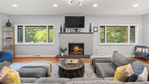 A modern living room with a fireplace, grey sofas, a coffee table, and large windows offering a view of greenery. - Simply Shores by AvantStay | Private Beach + Dock | Lake Overlook (Long Pond)