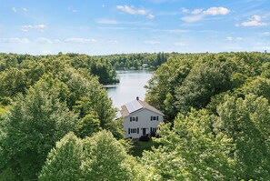 A two-story house with a gabled roof, surrounded by dense forest, and a lake in the background. - Lakefront Retreat • Hot Tub + Fire Pit + Private Beach | Simply Shores | (Long Pond)