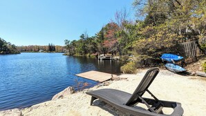 A beach chair on a sandy shore with a wooden dock extending into a lake, surrounded by trees and a fence. - Simply Shores by AvantStay | Private Beach + Dock | Lake Overlook (Long Pond)