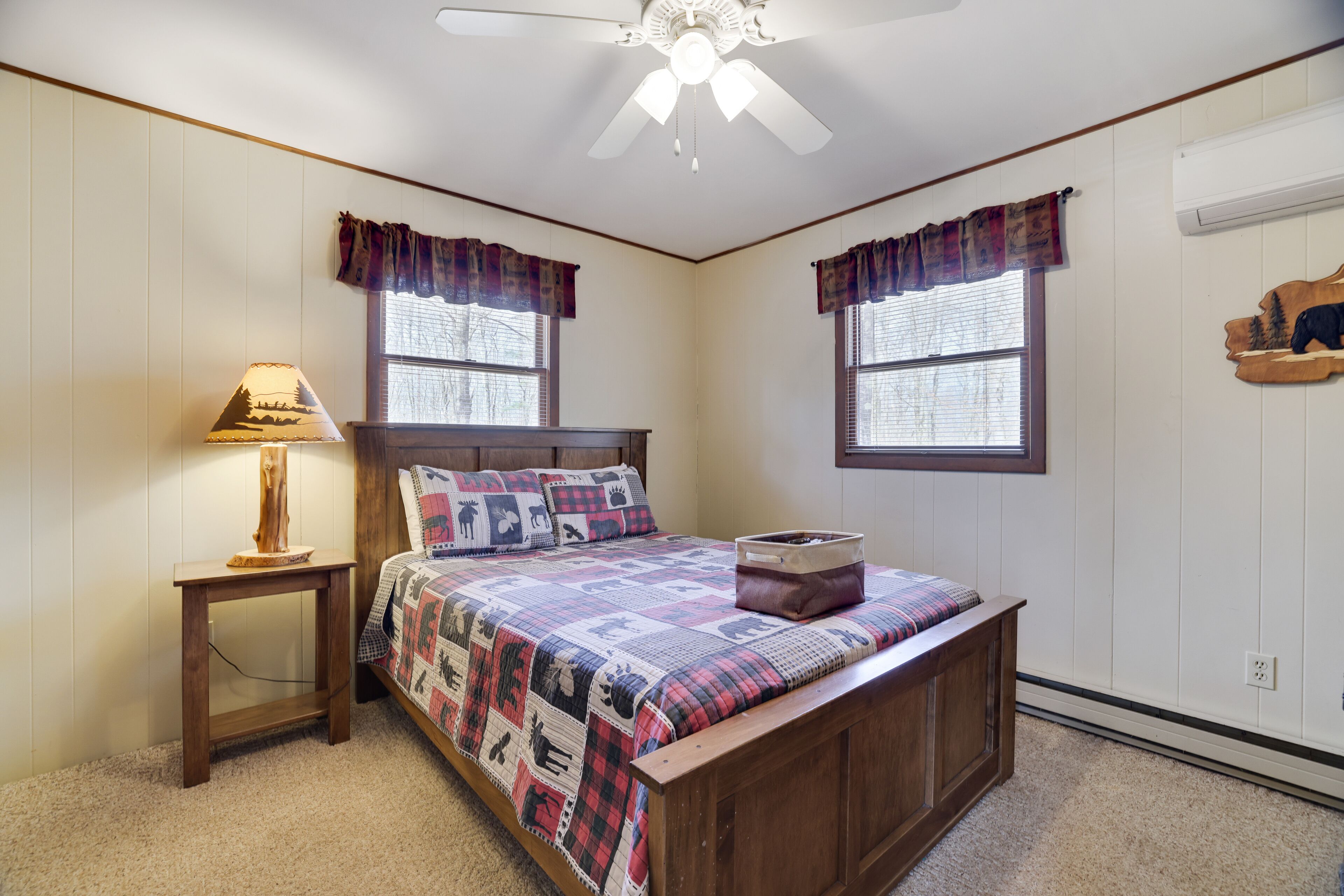 A bedroom with a wooden bed, a nightstand, a lamp, two windows with curtains, a ceiling fan, and a wall-mounted air conditioner.