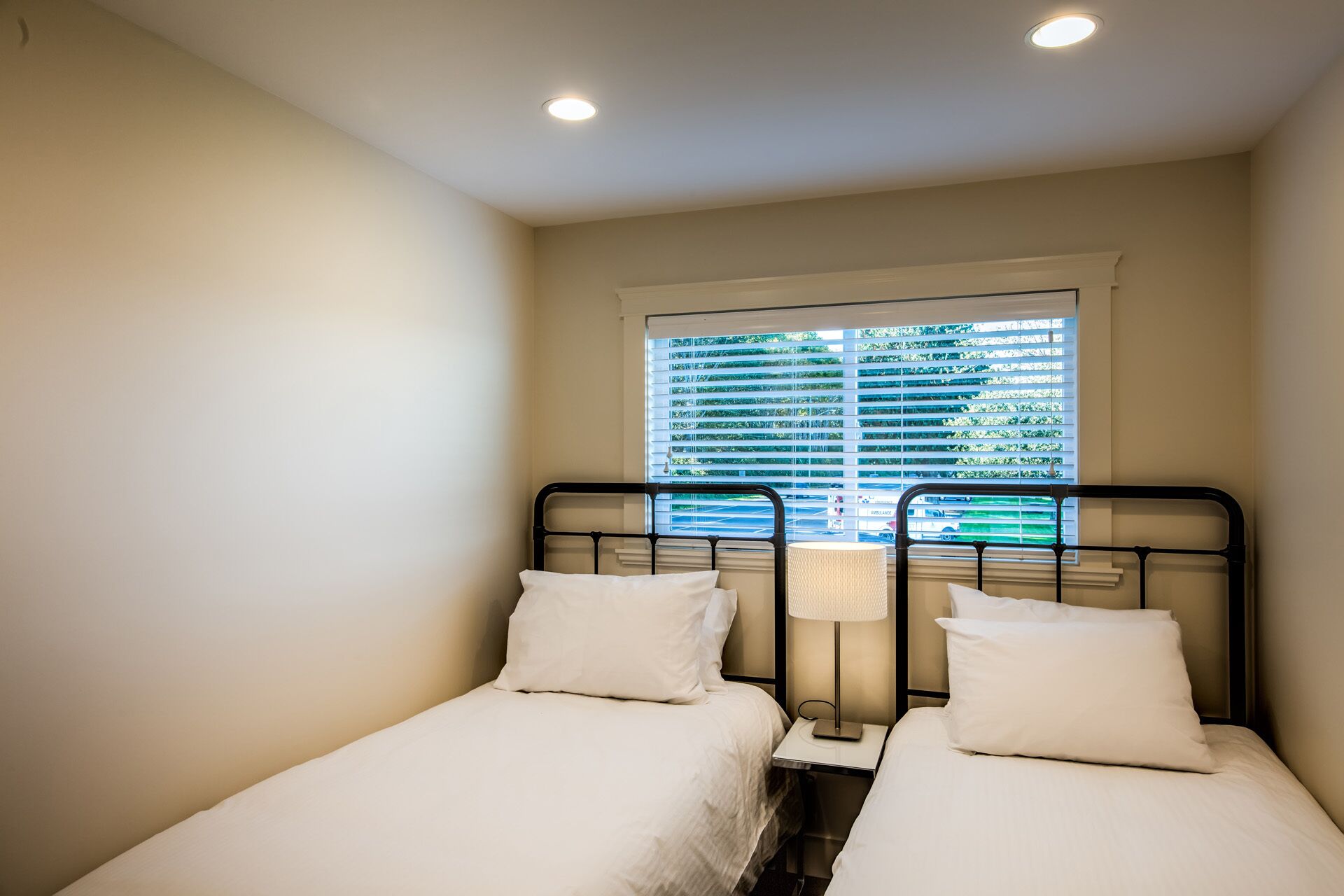 Una habitación pequeña y limpia con dos camas individuales, una ventana con persianas y una mesita de noche con lámpara.