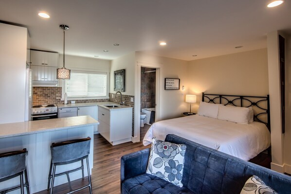 Une chambre d’hôtel de taille réduite comprenant un lit, une petite kitchenette, un coin salon avec un canapé et des fauteuils, et une salle de bain.