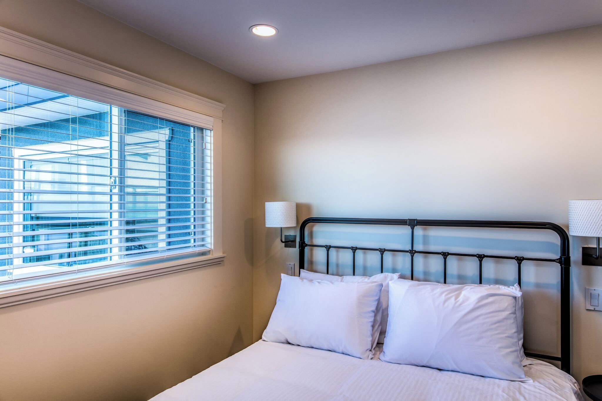 A bedroom with a bed, white bedding, a window with blinds, and a wall-mounted lamp.